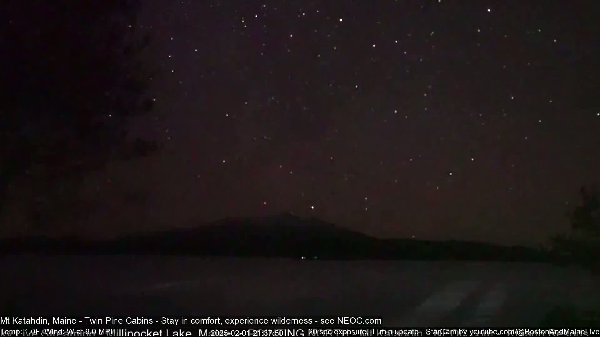 A dark, star-filled night sky overlooks a calm body of water with a silhouetted mountain in the distance.