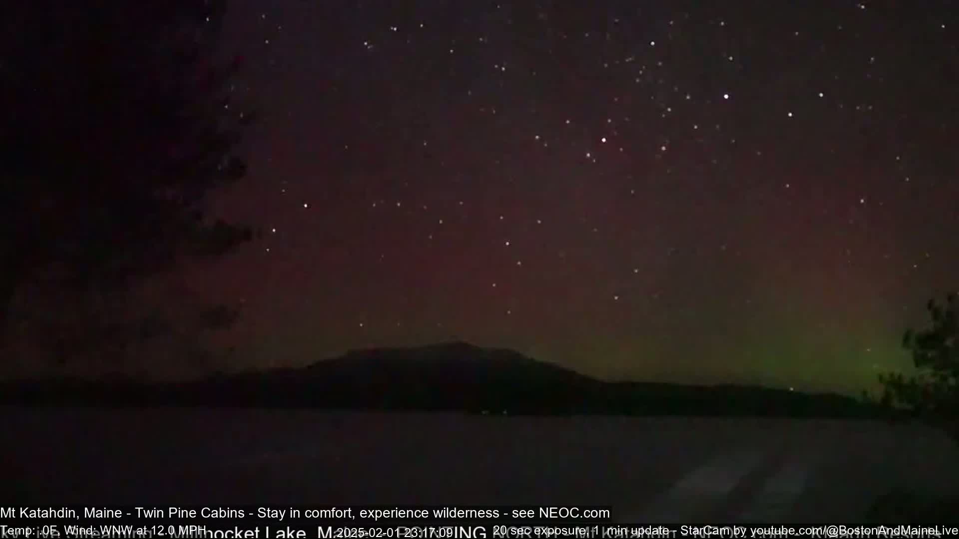 A dark night sky filled with stars and a faint aurora borealis displays over a silhouetted mountain and a dark body of water.