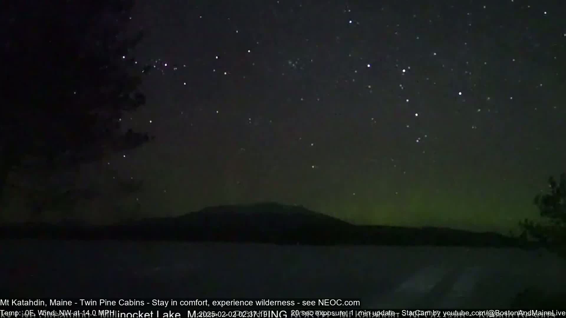 A nighttime view of a dark, starry sky with a faint green aurora borealis over a silhouetted mountain and a lake.
