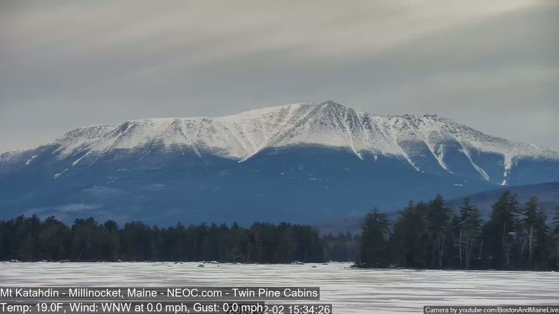A snow-covered mountain rises behind a dark evergreen forest and a partially frozen lake under a cloudy sky.