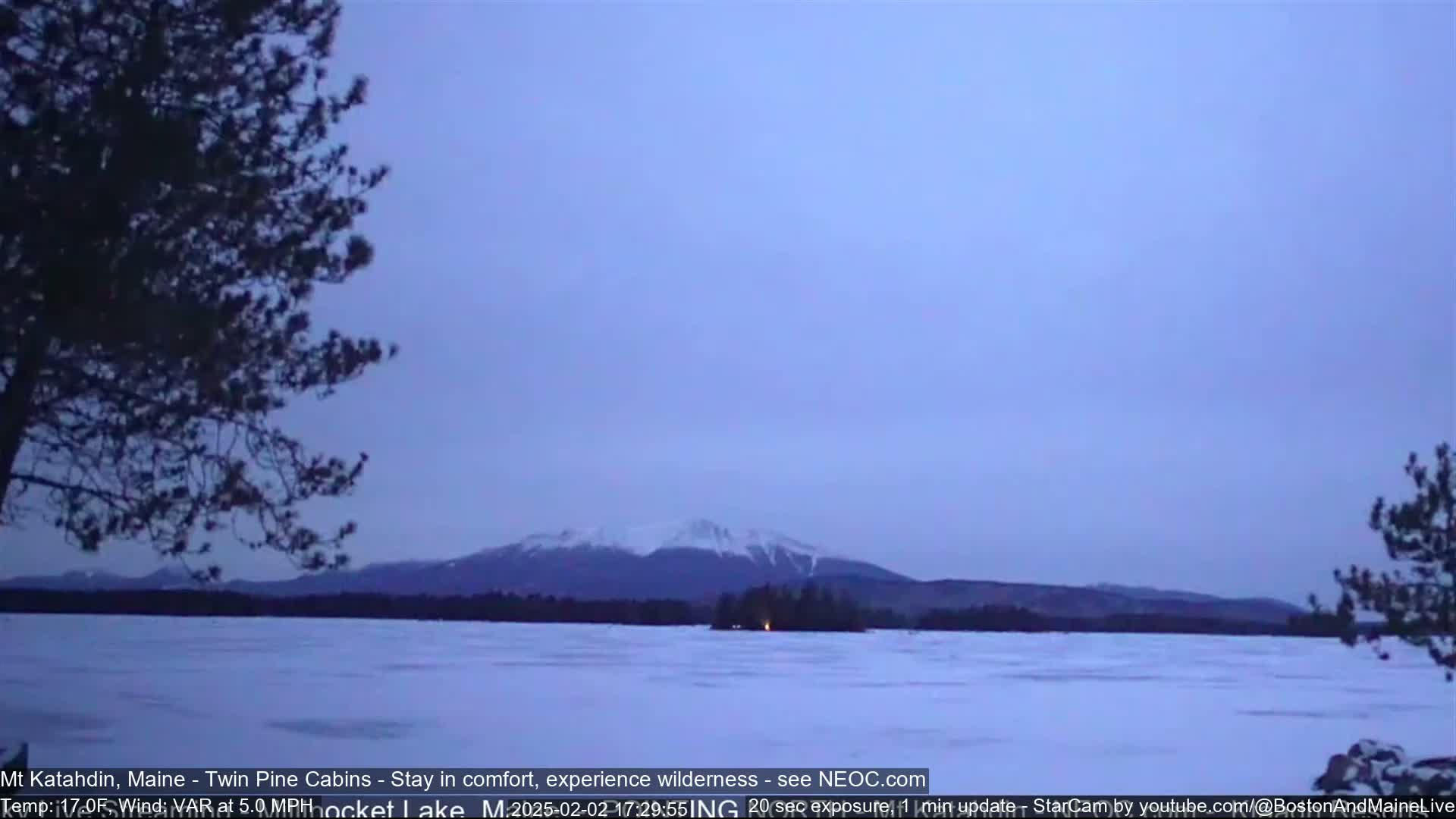 A snow-covered lake stretches towards a distant, snow-capped mountain range under a dusky sky with a light breeze.