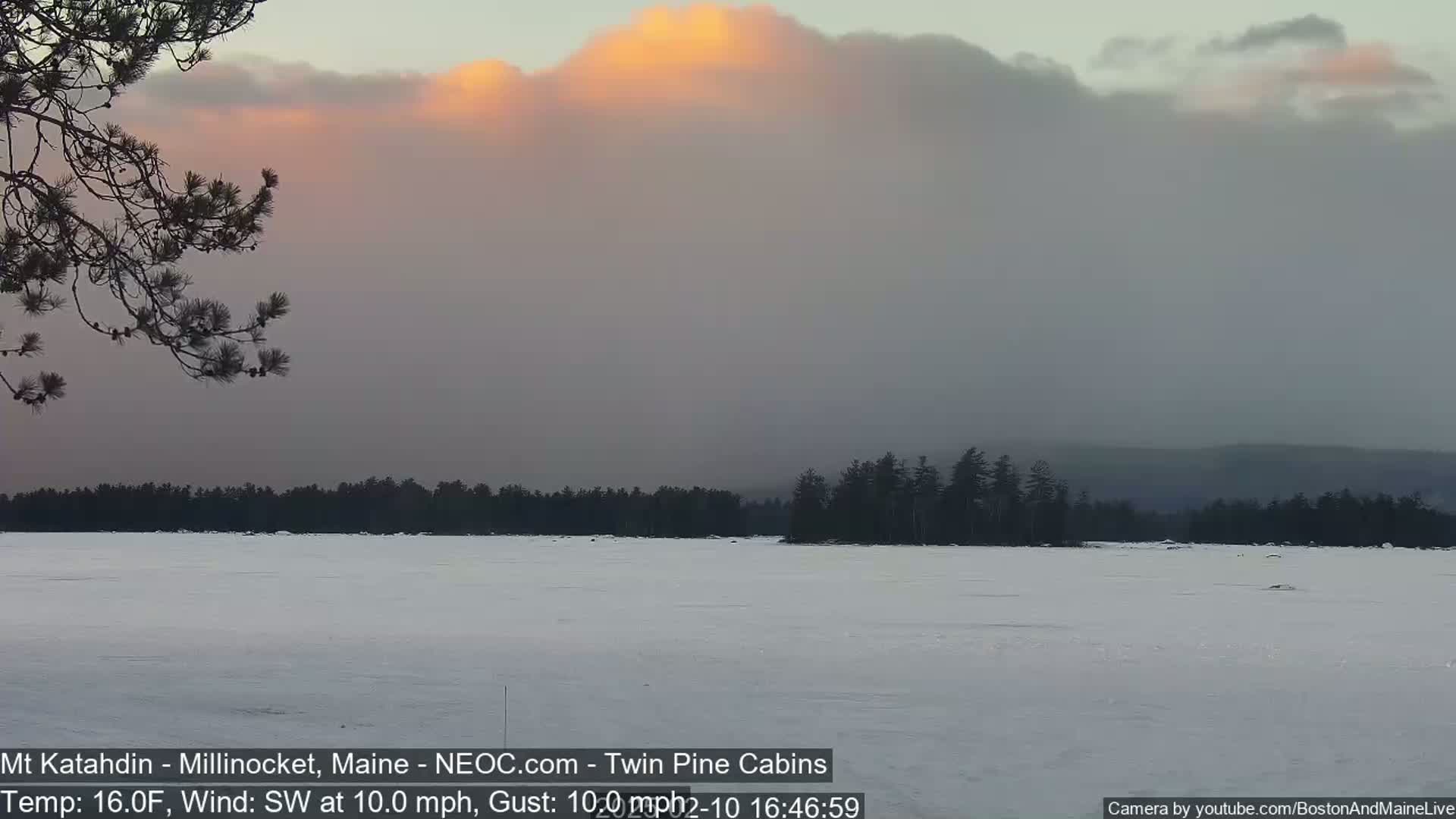 A snow-covered frozen lake stretches to a dark treeline under a cloudy sky with a patch of orange-tinged clouds near the horizon.