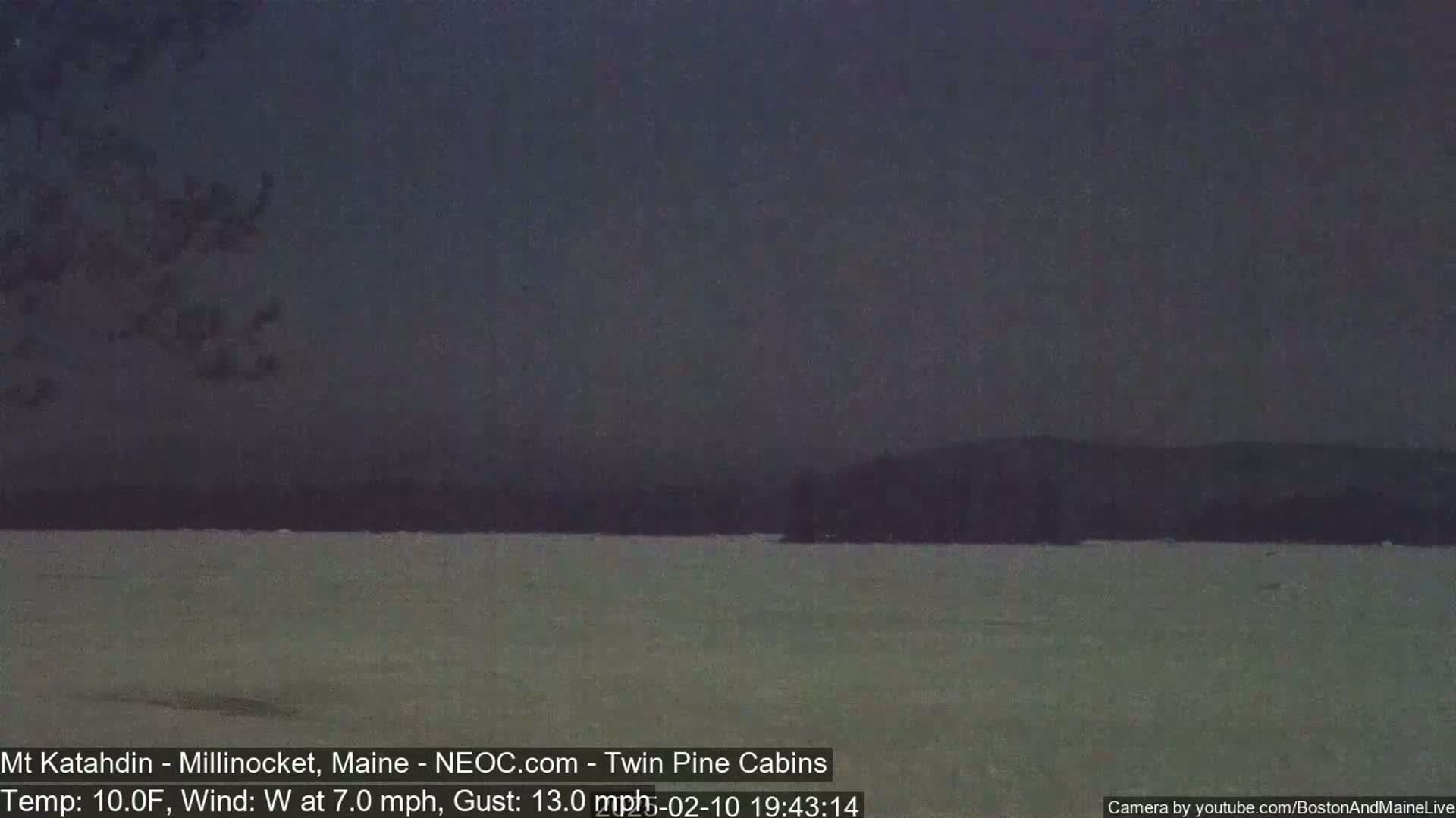 A dark, overcast view of a snow-covered lake with a distant, silhouetted treeline under a dark sky.