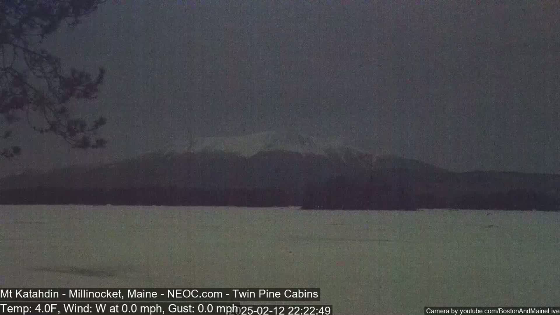 A snow-covered mountain range is visible in the distance across a calm, snow-covered lake under a dark, overcast sky.