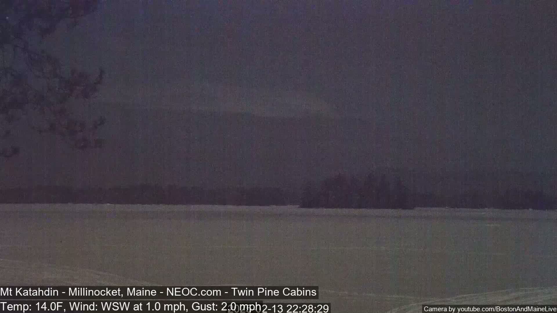 A dark, snowy landscape shows a body of water in the foreground, a treeline in the middle ground, and distant mountains under a dusky sky.
