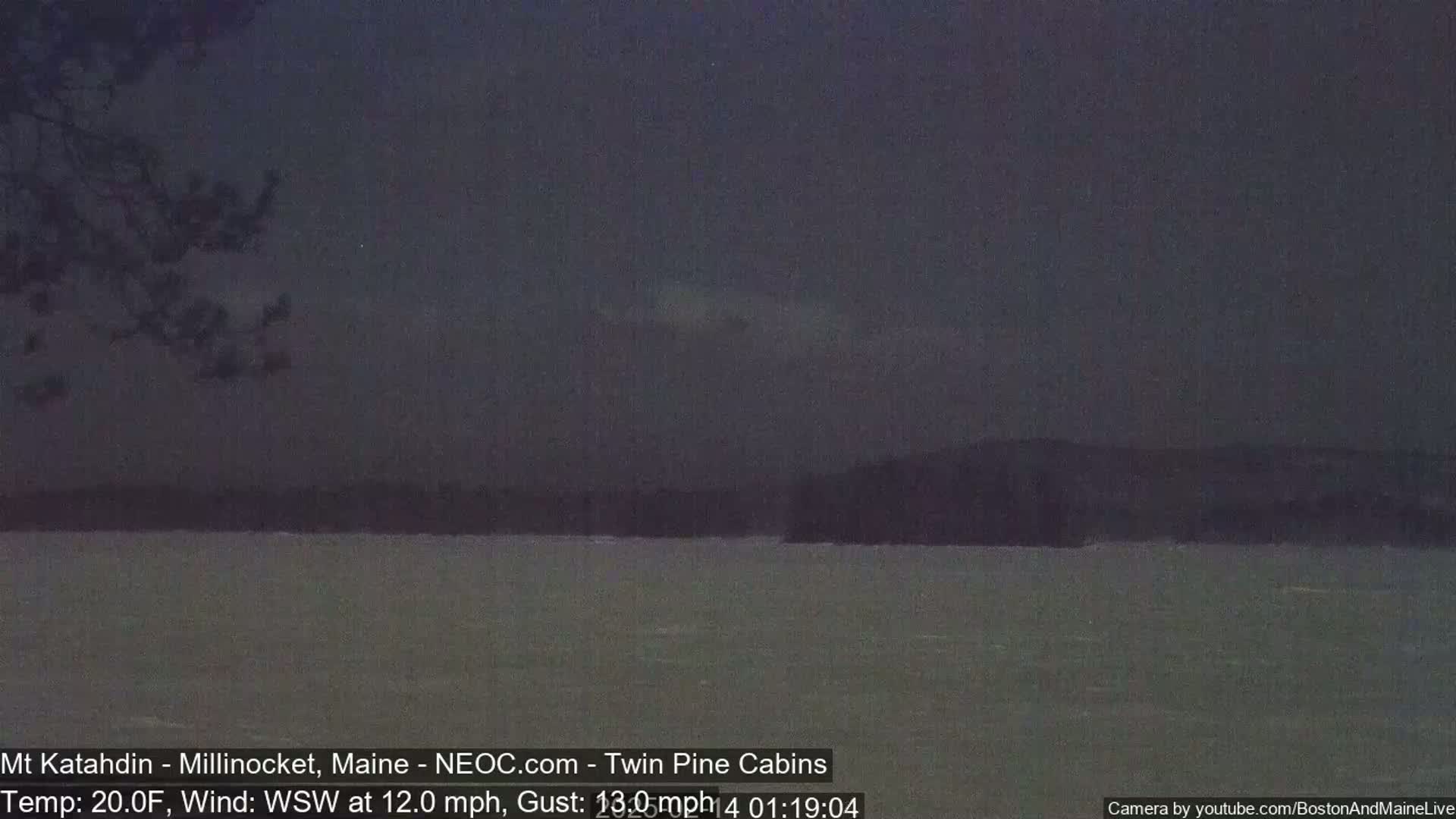 A dark, nighttime view of a calm body of water with a distant, dark shoreline under a cloudy sky.
