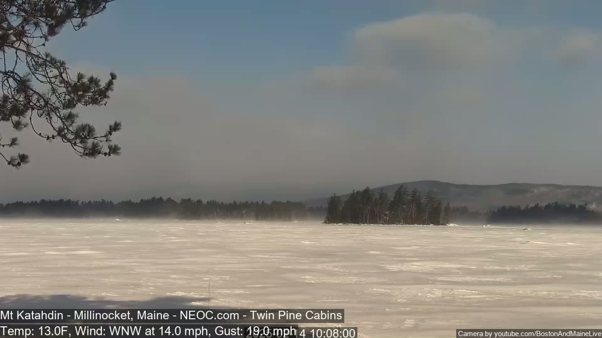 A snow-covered, frozen lake stretches to a distant, wooded shoreline under a partly cloudy sky with a 13°F temperature and a WNW wind at 14 mph gusting to 19 mph.