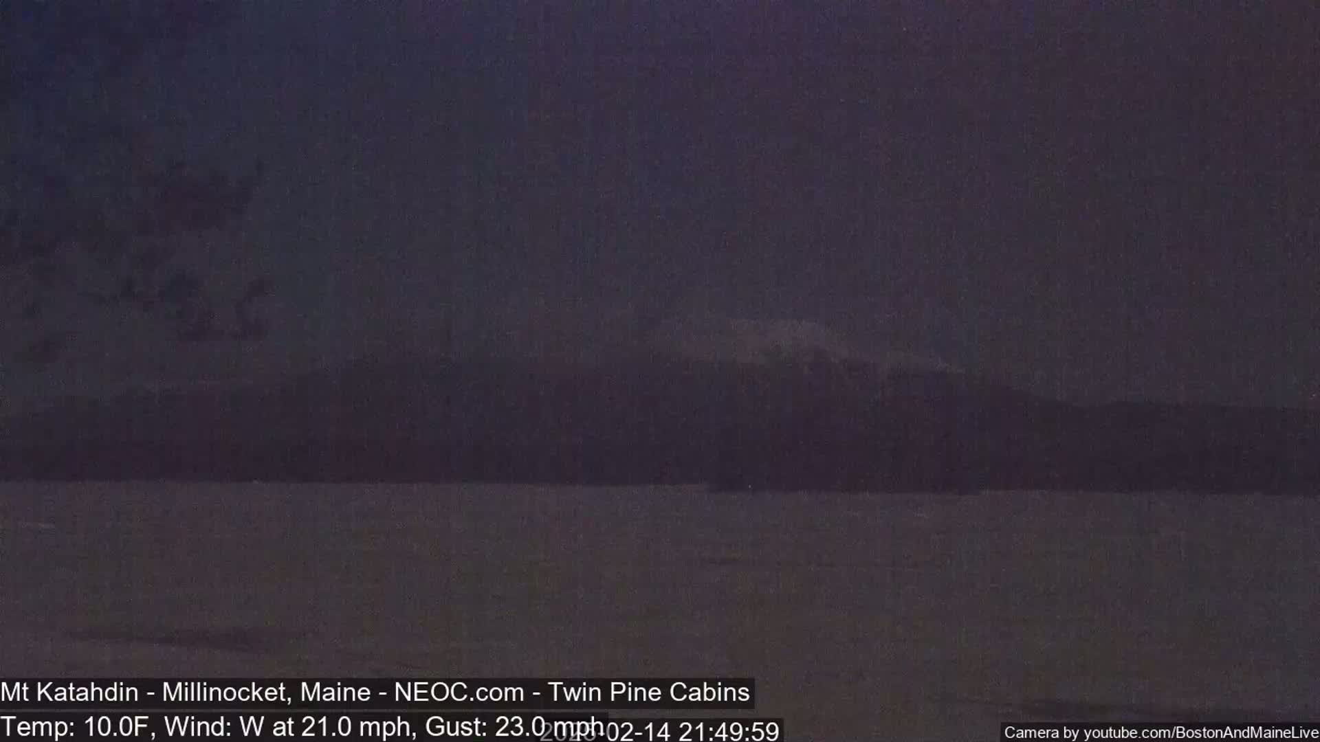 A dark, nighttime view of a calm body of water with a mountain range in the background under a cloudy sky.