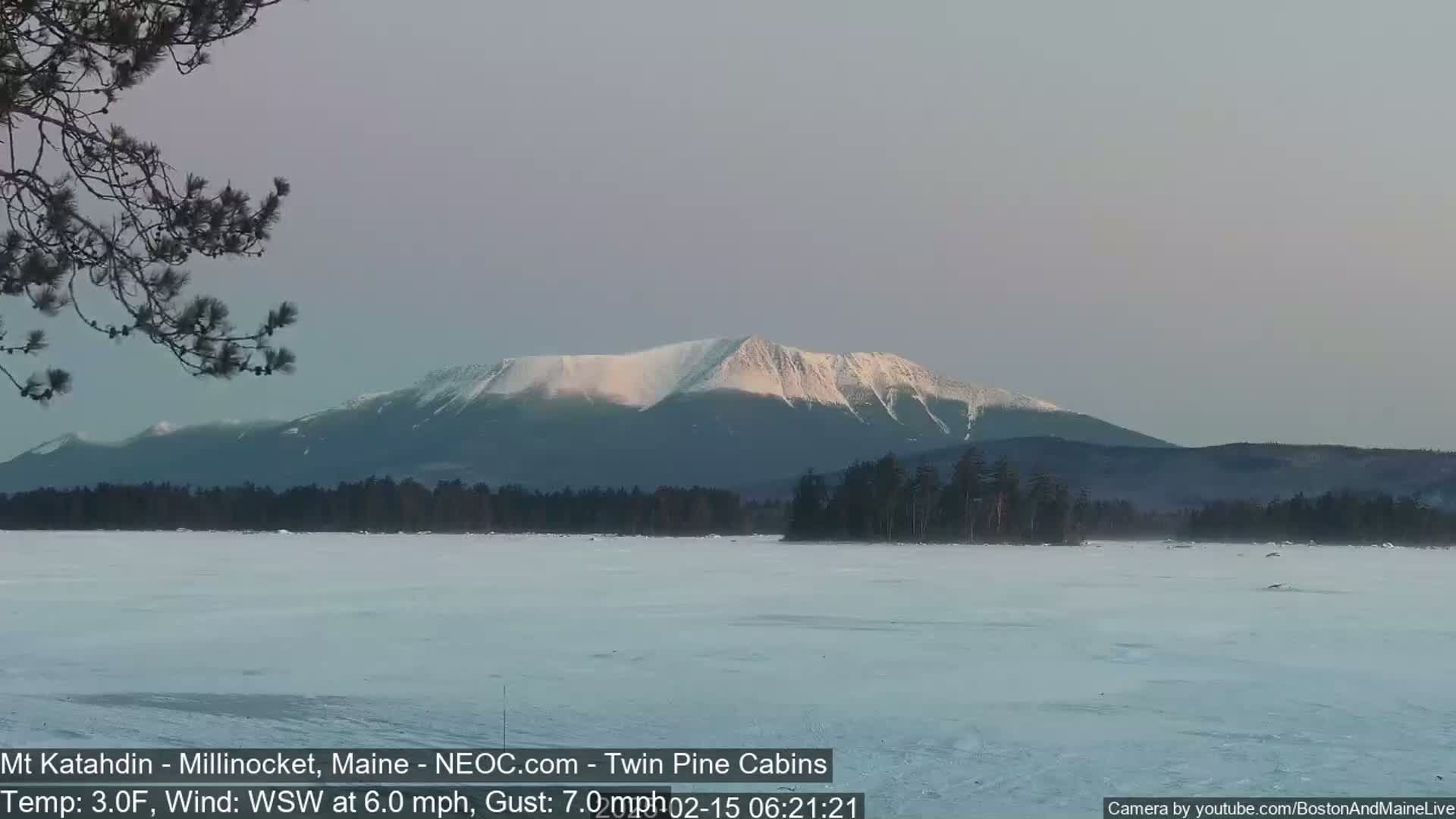 A snow-covered lake stretches out to a snow-capped mountain range under a pale sky, with a light wind blowing.