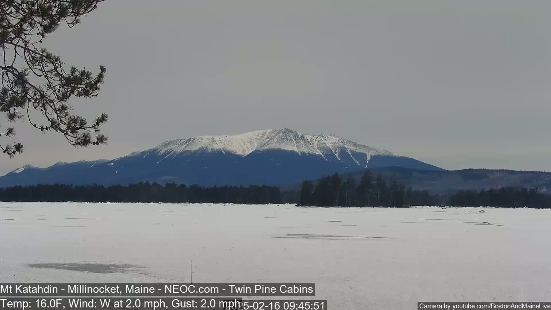 A snow-covered, frozen lake extends to a background of snow-capped mountains under a gray sky, with a gentle westerly wind.