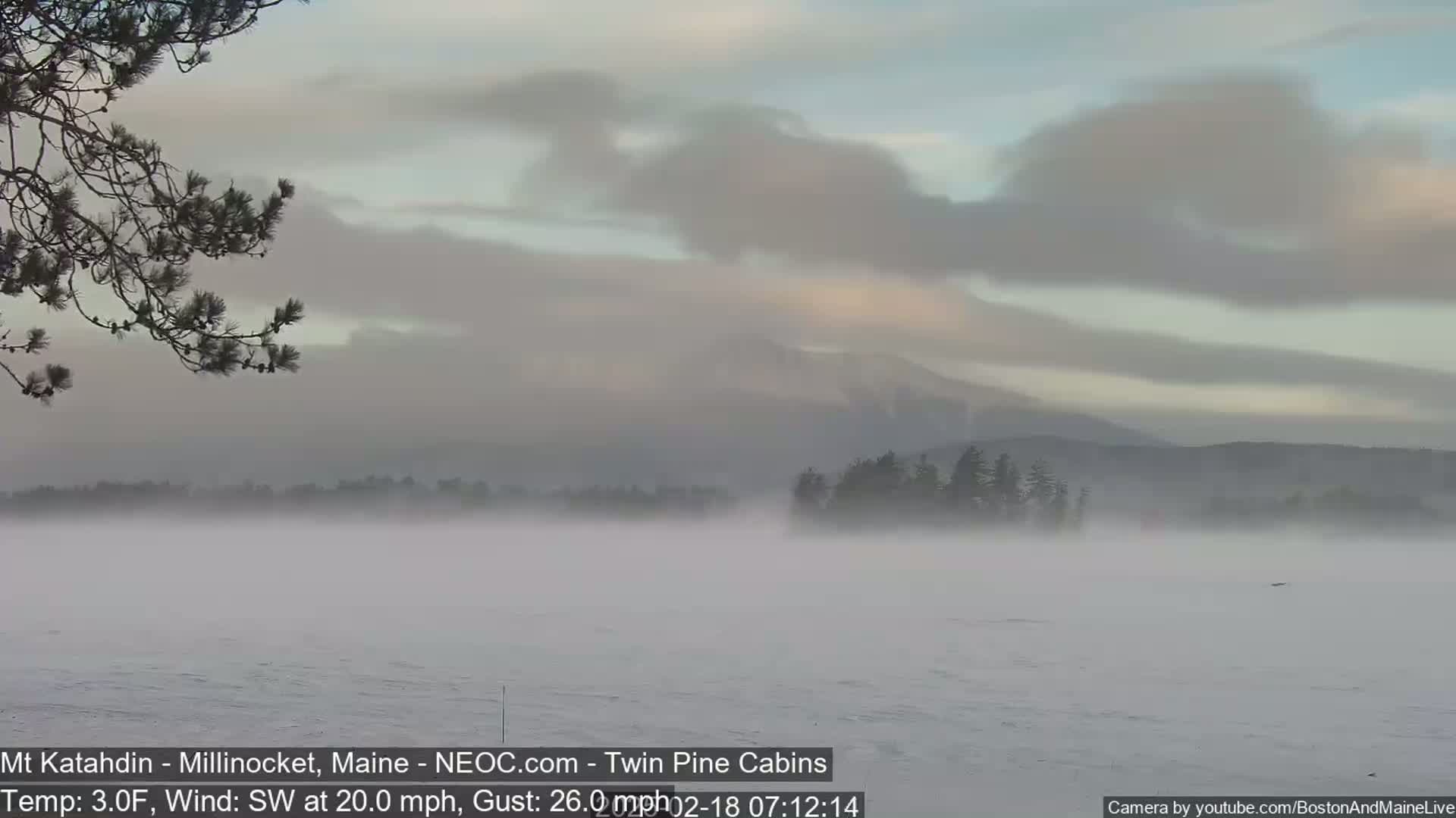 A foggy, snowy landscape shows a calm body of water with distant mountains and trees under a cloudy sky.