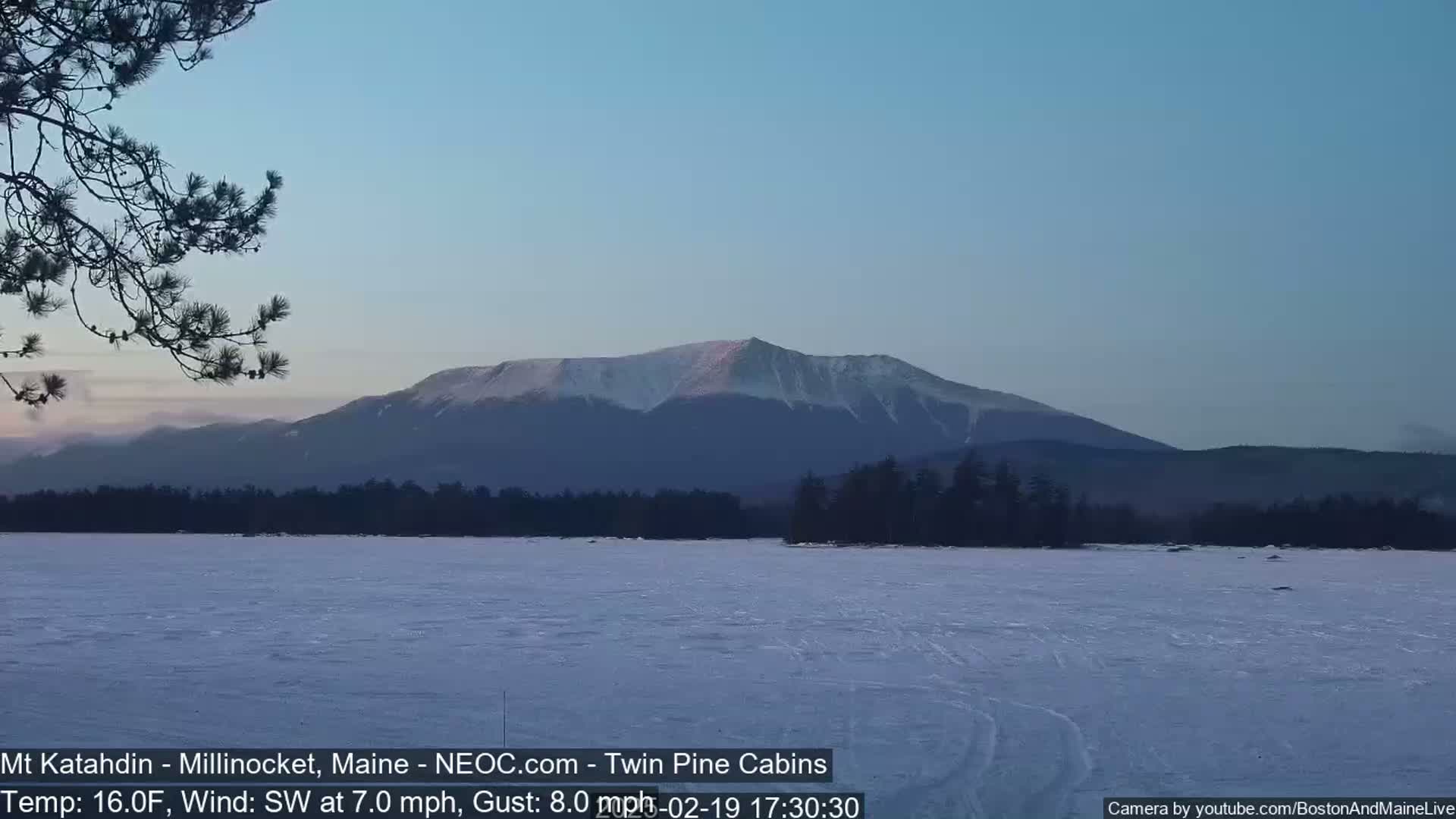 A snow-covered lake stretches before a snow-capped mountain range under a clear, twilight sky with a southwest wind.