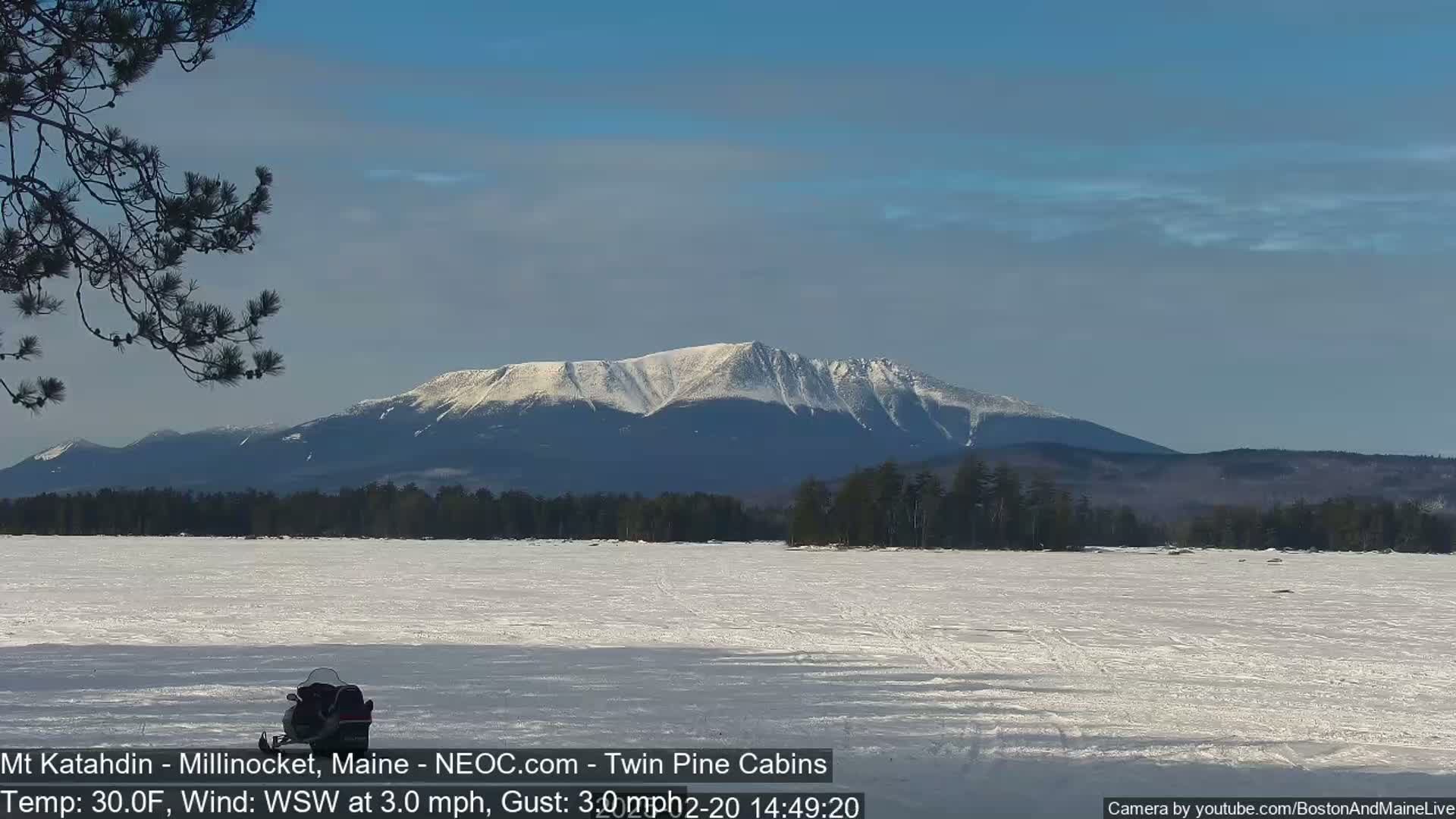 A snowmobile sits on a frozen lake in front of a snow-covered mountain range under a clear blue sky.