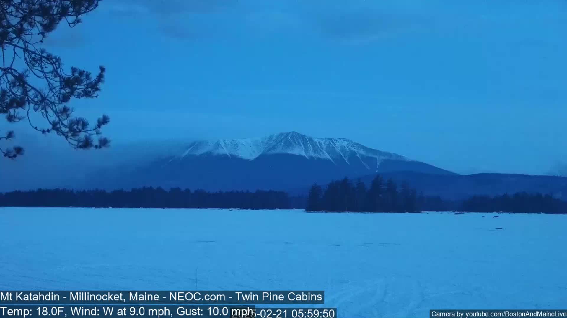 A snow-covered lake stretches out before a snow-capped mountain range under a twilight blue sky, with a light westerly wind.