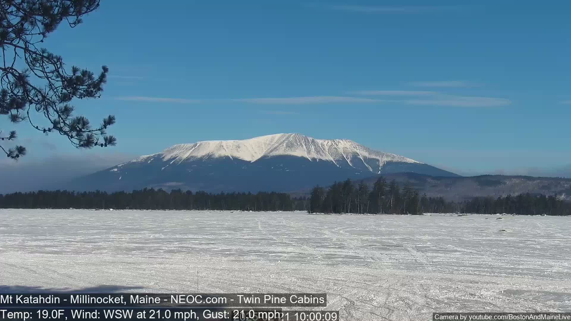 A snow-covered, frozen lake stretches to a background of a snow-capped mountain range under a clear blue sky, with a strong wind blowing from the west-southwest.