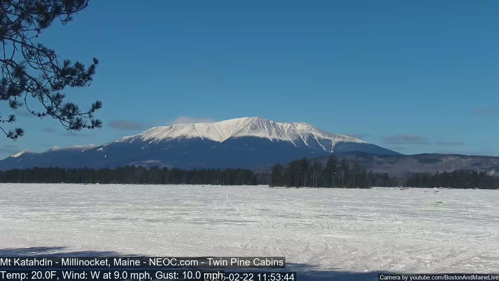 A snow-covered lake stretches to a snow-capped mountain range under a clear blue sky, with a gentle westerly wind.