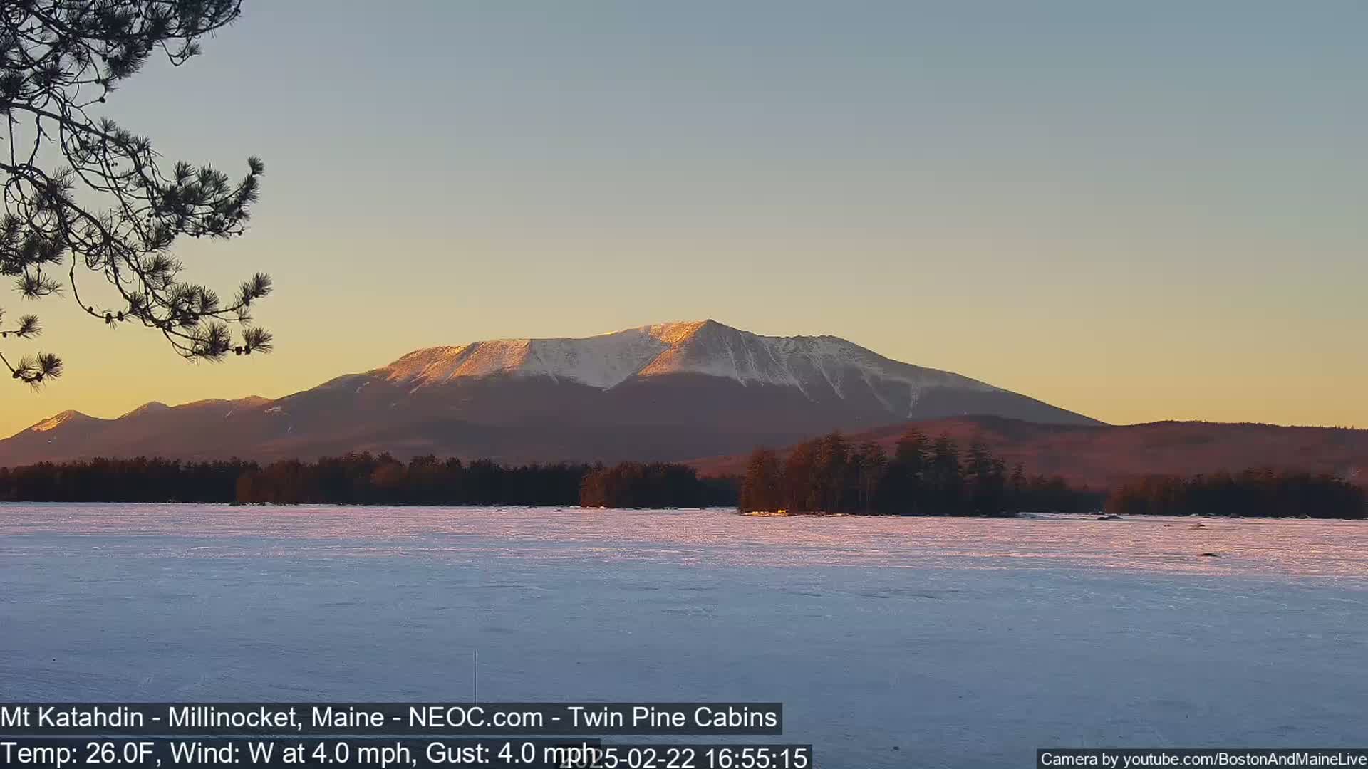 A snow-covered lake stretches before a snow-capped mountain range at sunset under a clear sky.