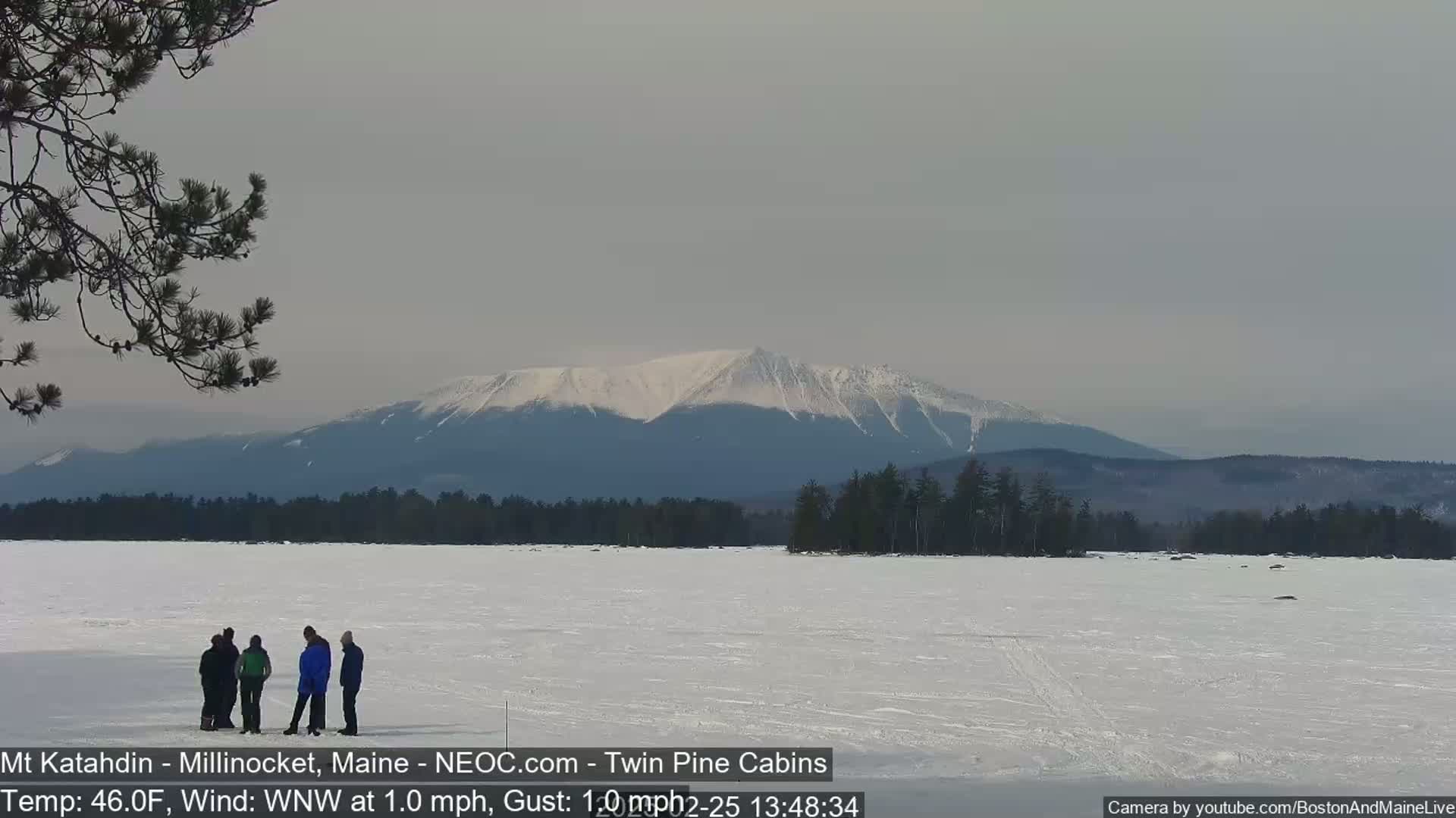 A group of people stand on a frozen lake with a snow-covered mountain range in the background under a cloudy sky.