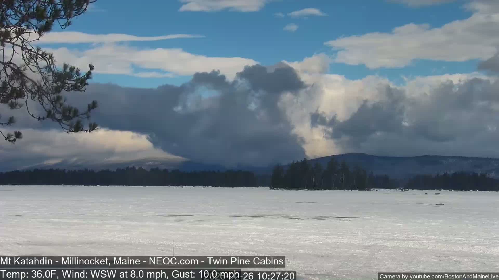 A partially frozen lake stretches to a distant, tree-covered mountain range under a partly cloudy sky with a strong wind.