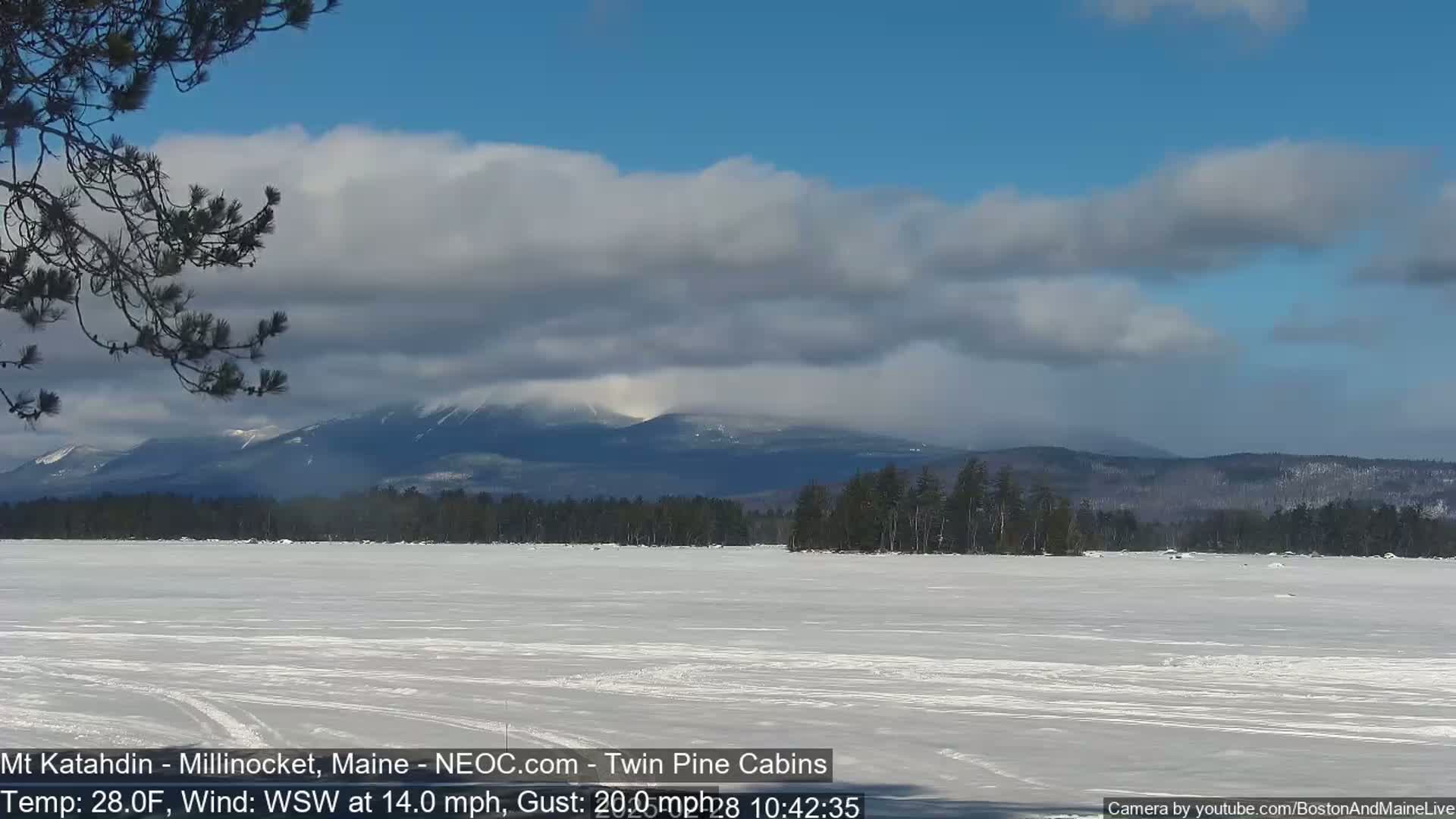 A snow-covered lake stretches to a background of distant, snow-capped mountains under a partly cloudy sky, with a wind blowing from the west-southwest at 14 mph and gusts up to 20 mph.