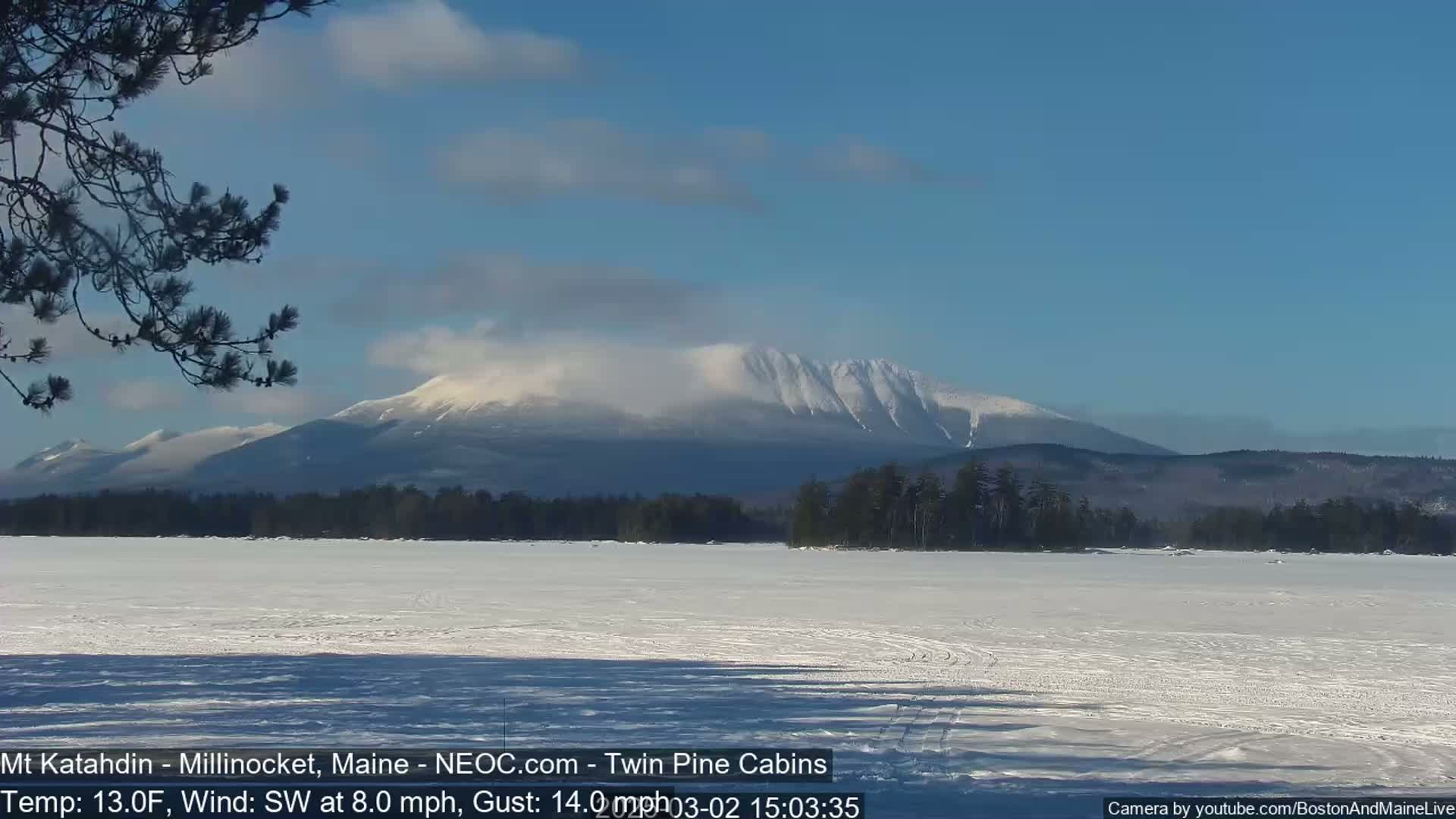 A snow-covered frozen lake stretches out before a snow-capped mountain range under a mostly clear blue sky, with a light southwest wind.