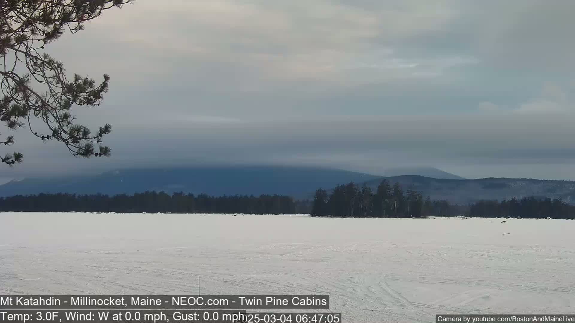 A snow-covered frozen lake stretches to a background of dark, distant mountains under a mostly cloudy, gray sky.