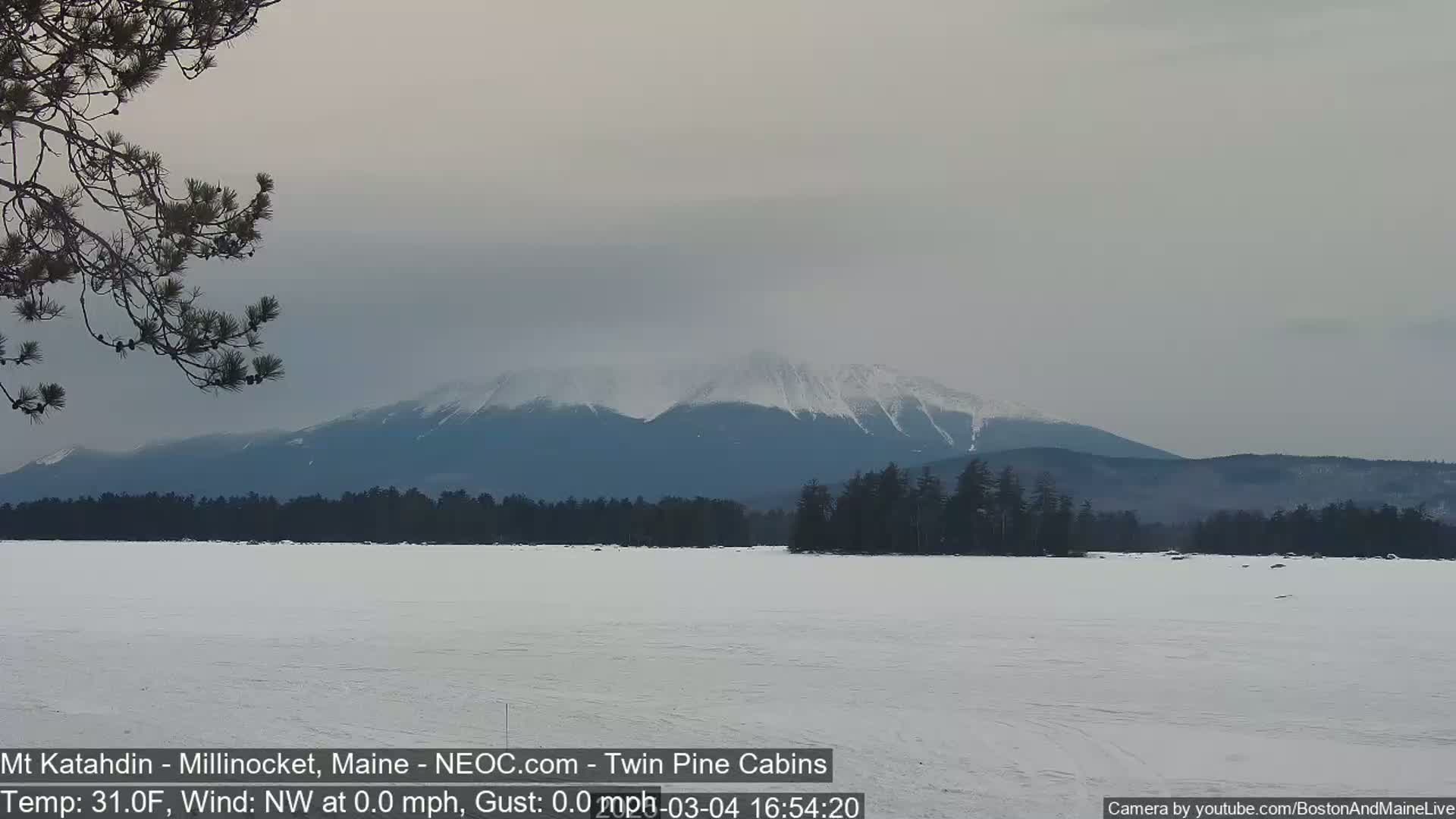 A snow-covered, frozen lake stretches out before a range of snow-capped mountains under an overcast sky.