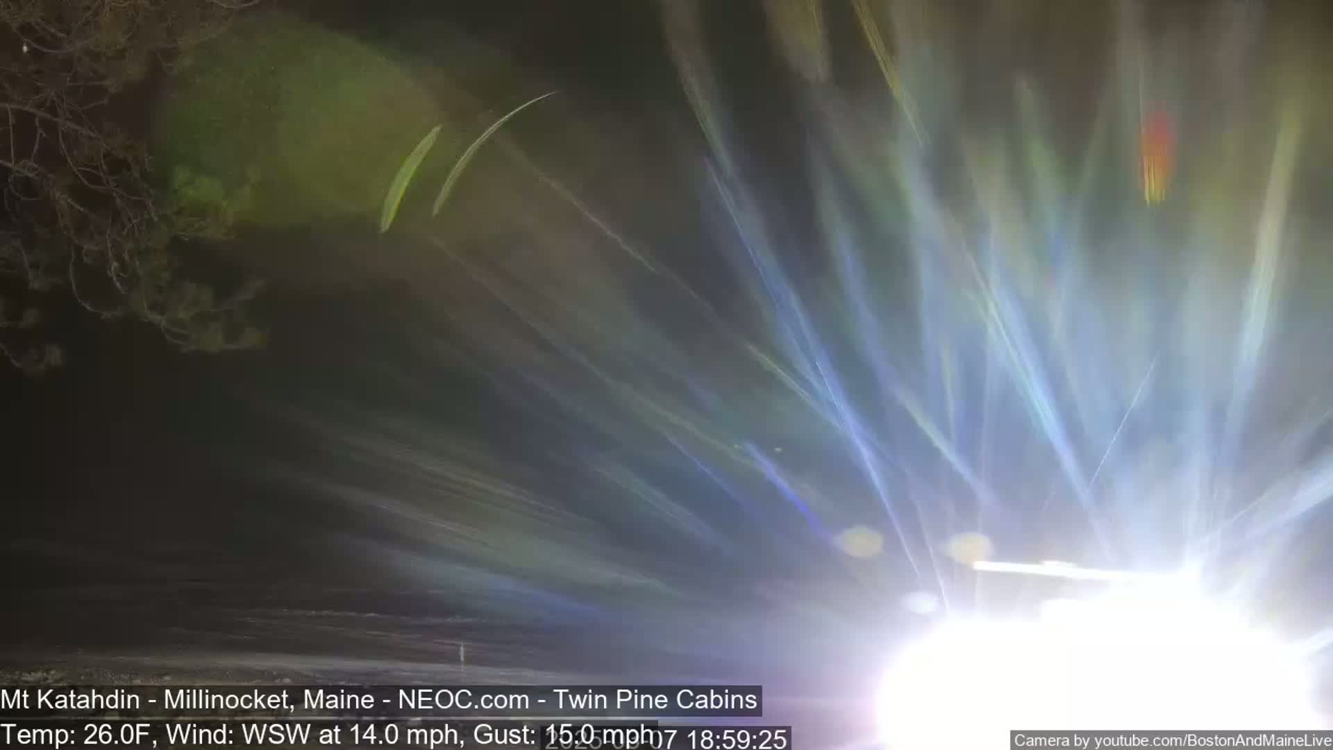 A nighttime, blurry image shows bright streaks of light emanating from a source at the bottom of the frame, against a dark background of trees and a 26°F temperature with a 14 mph wind.