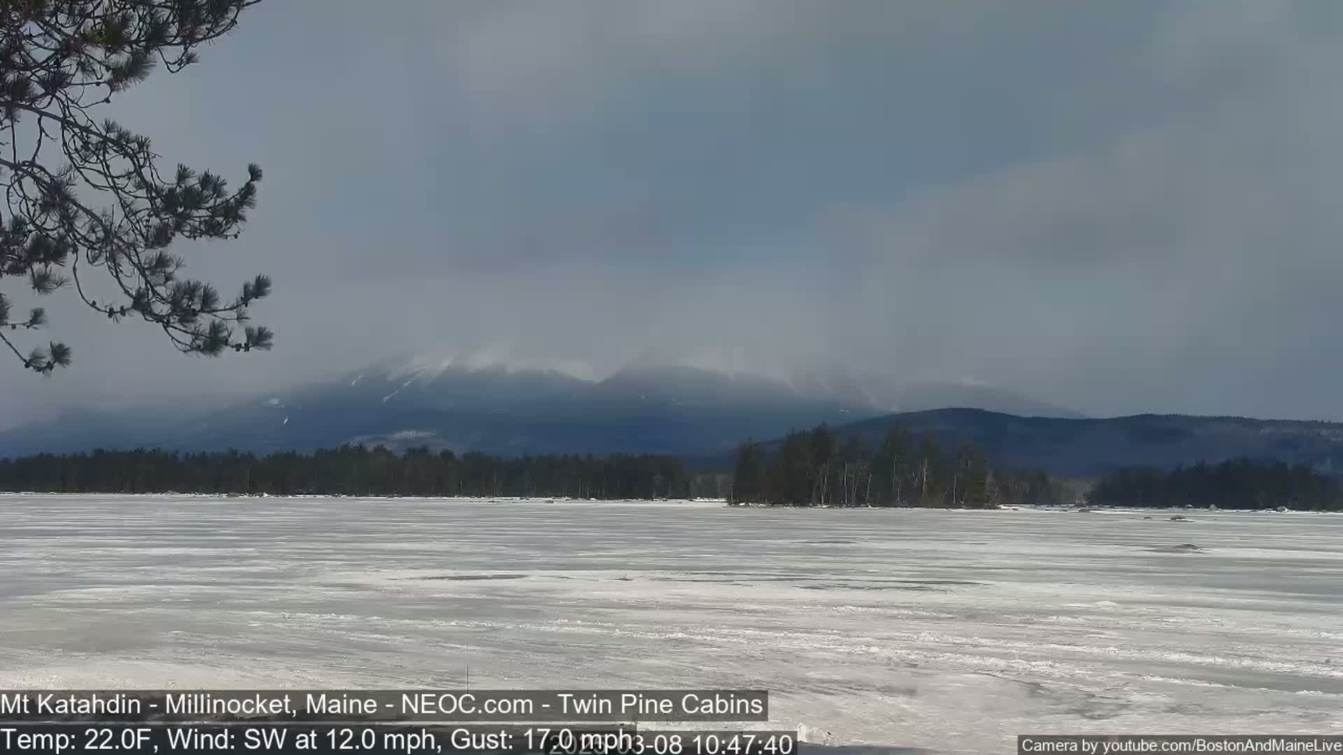 A partially frozen lake stretches towards snow-covered mountains under a mostly cloudy sky.