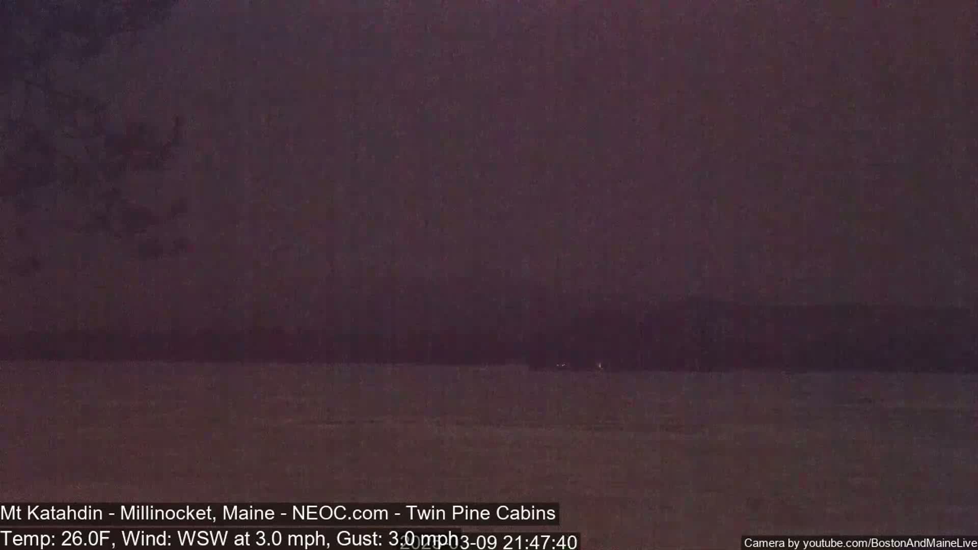 A dark, nighttime view of a calm body of water with a dark shoreline and trees in the distance under a dusky sky.