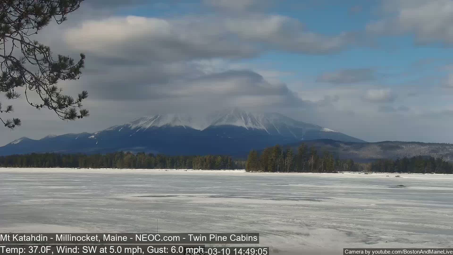 A partially frozen lake stretches before snow-capped mountains under a partly cloudy sky.