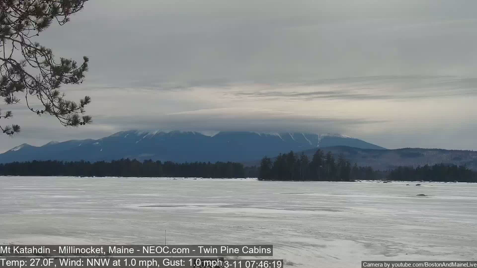 A frozen lake stretches to a snow-dusted mountain range under a mostly cloudy sky.