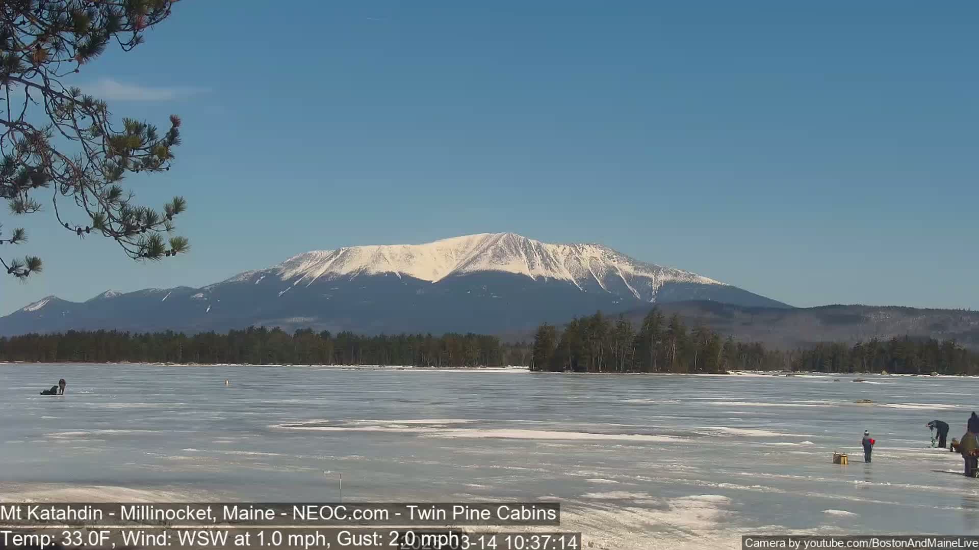 A snow-capped mountain range is visible in the distance across a frozen lake where several people are ice fishing on a clear, sunny day.