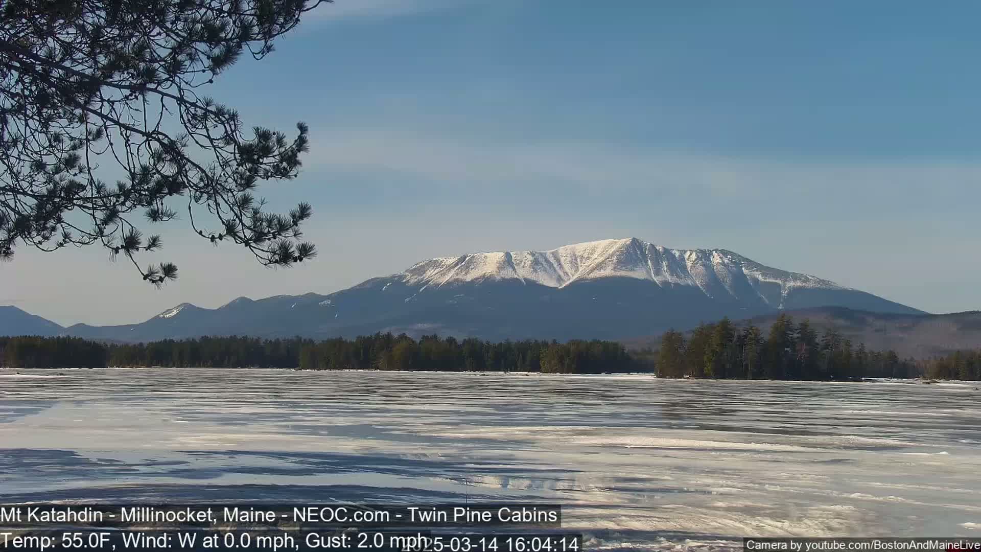 A partially frozen lake stretches out before a snow-capped mountain range under a clear blue sky.