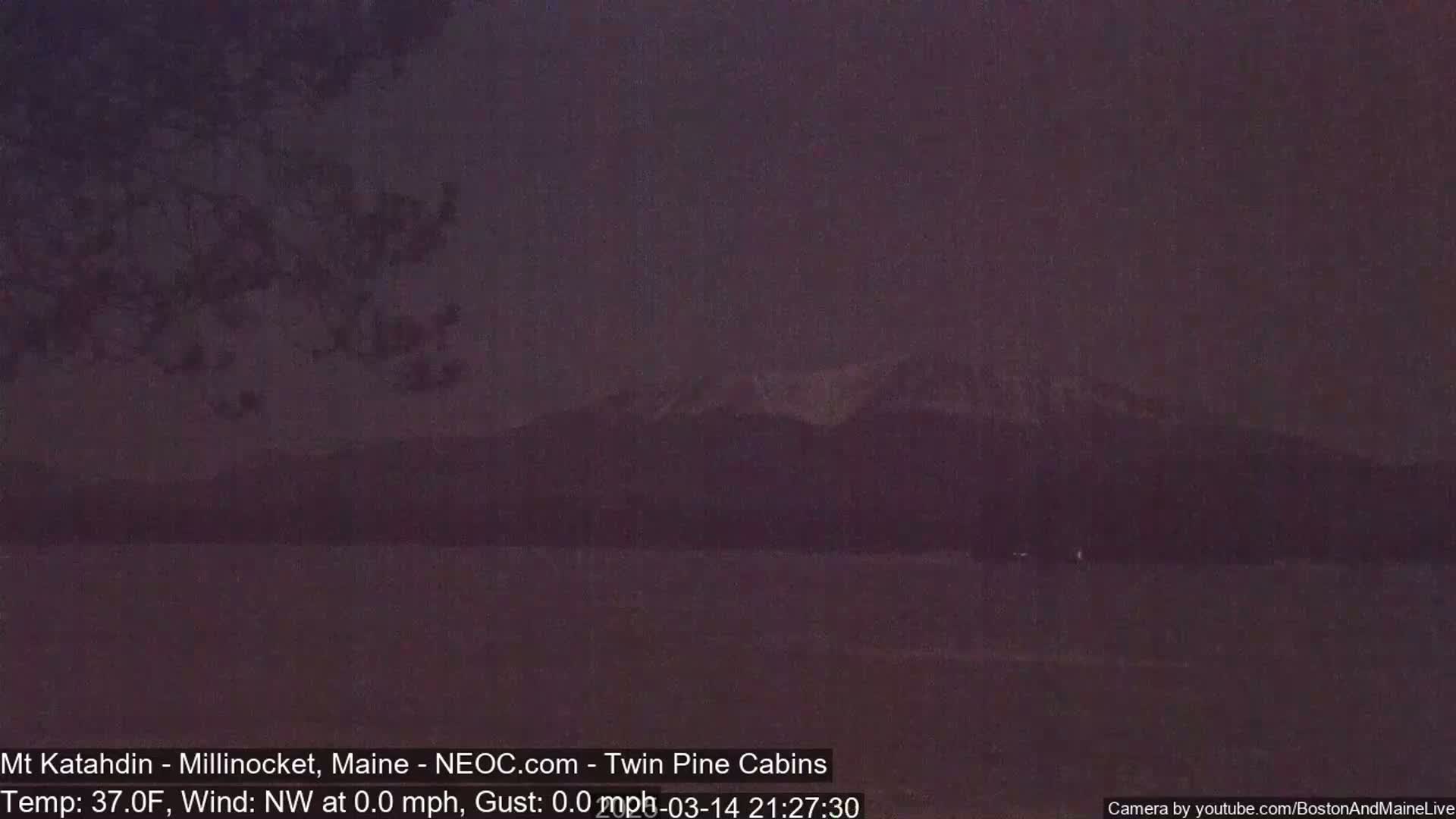 A dark, nighttime view of a snow-capped mountain range across a calm body of water under a dusky sky.