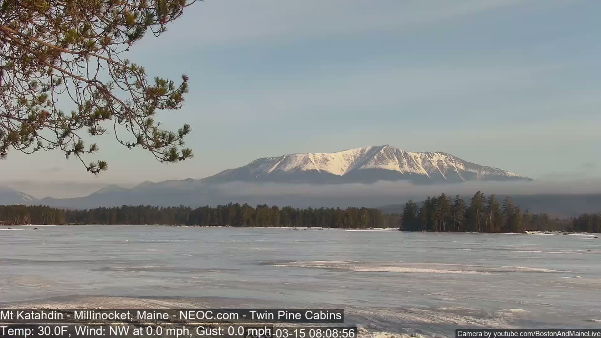 A snow-capped mountain range is visible across a partially frozen lake under a clear, calm sky.
