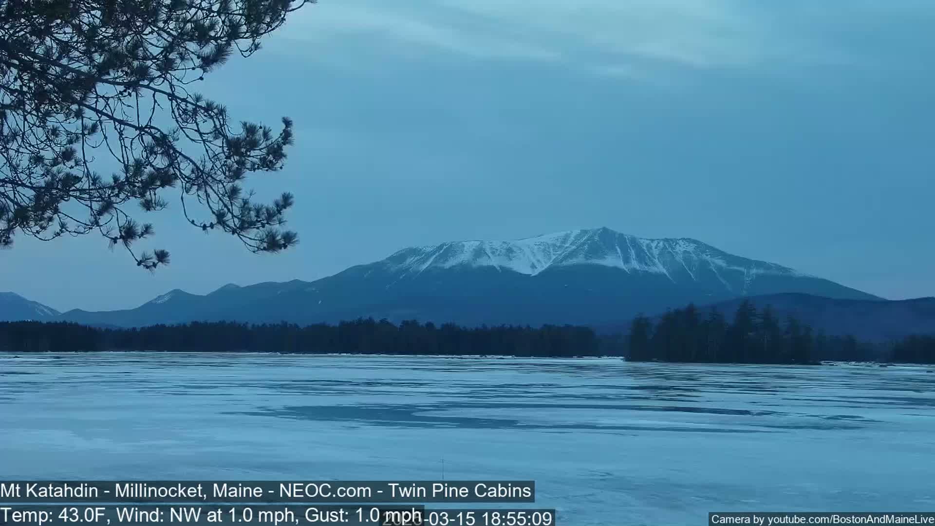 A partially frozen lake reflects a snow-capped mountain range under a twilight sky.