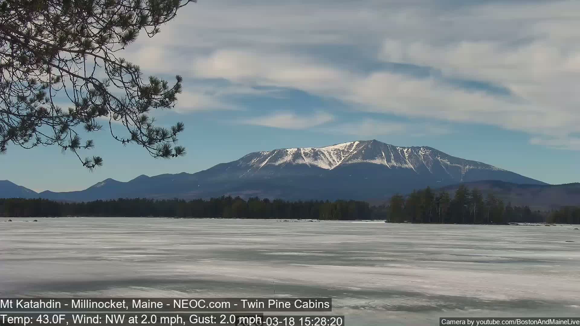 A partially frozen lake stretches towards a snow-capped mountain range under a partly cloudy sky with a light northwest wind.
