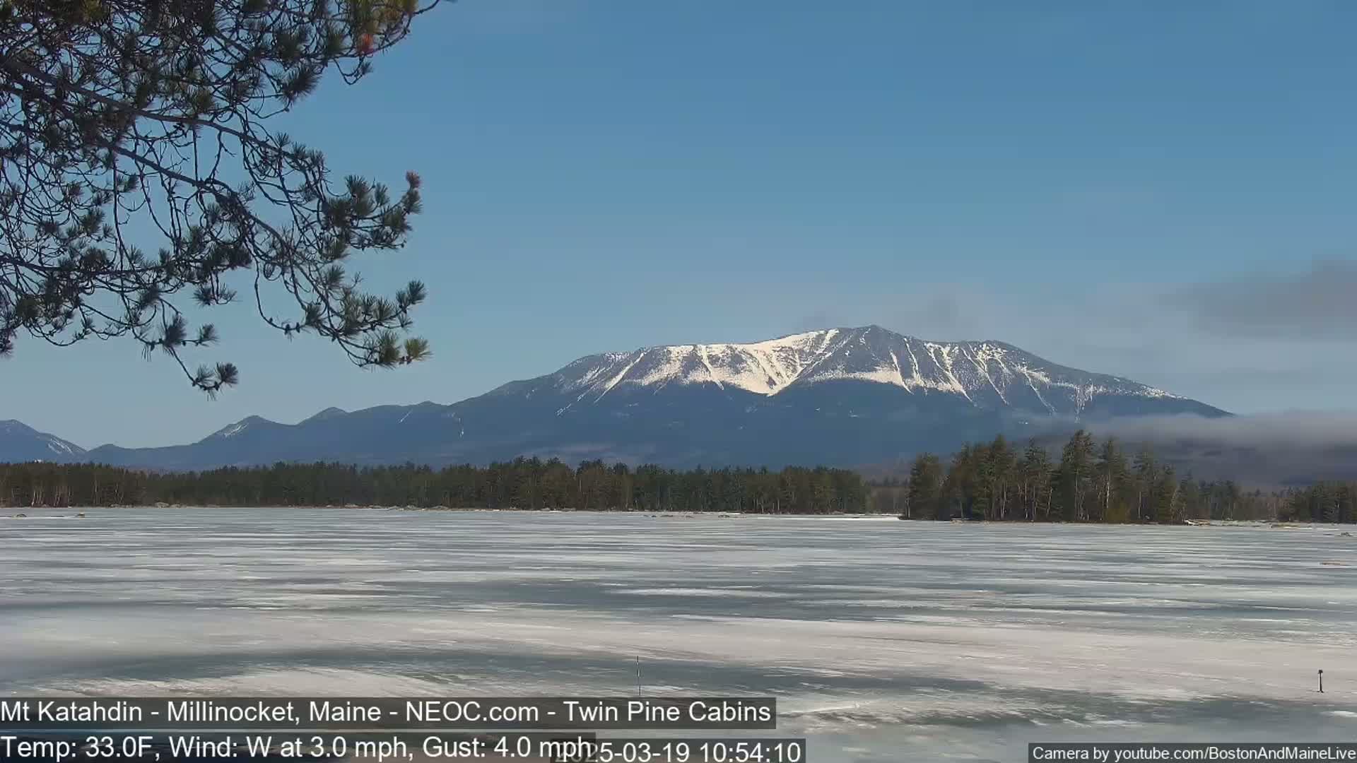 A partially frozen lake stretches to a snow-capped mountain range under a clear blue sky.