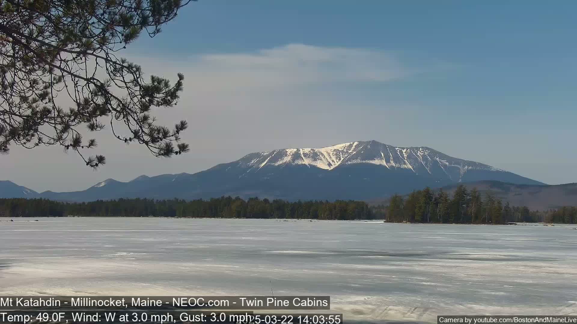 A partially frozen lake stretches out before a snow-capped mountain range under a mostly clear sky.