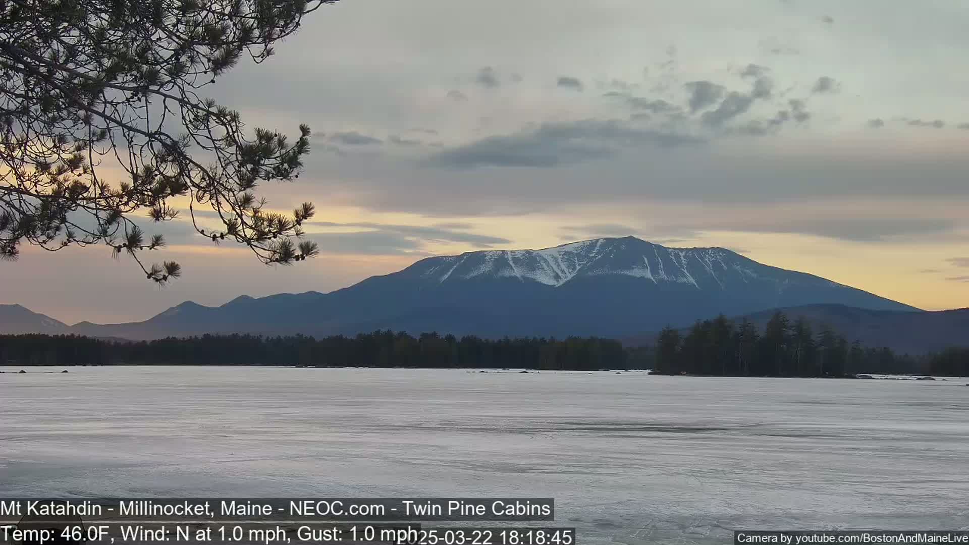 A partly cloudy sunset illuminates a snow-capped mountain range across a frozen lake, with a calm north wind.