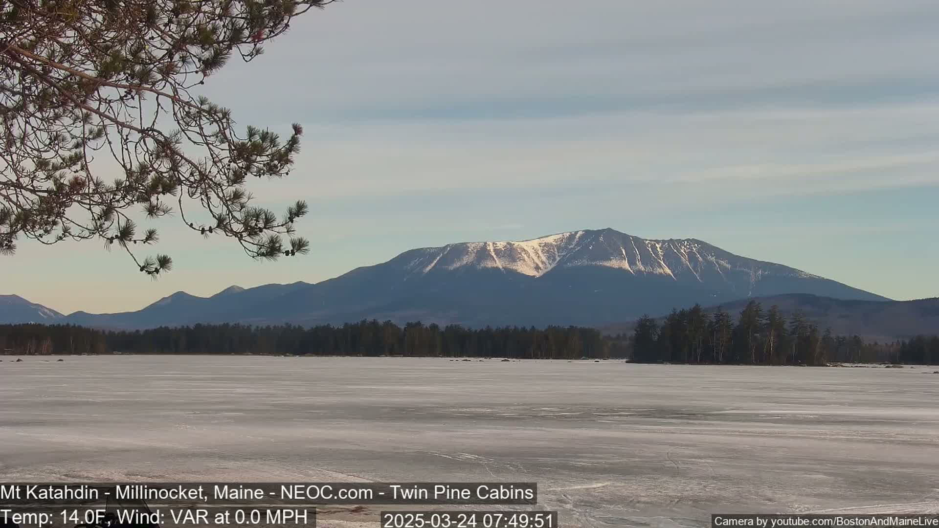 A partially frozen lake stretches out before a snow-capped mountain range under a mostly clear sky.