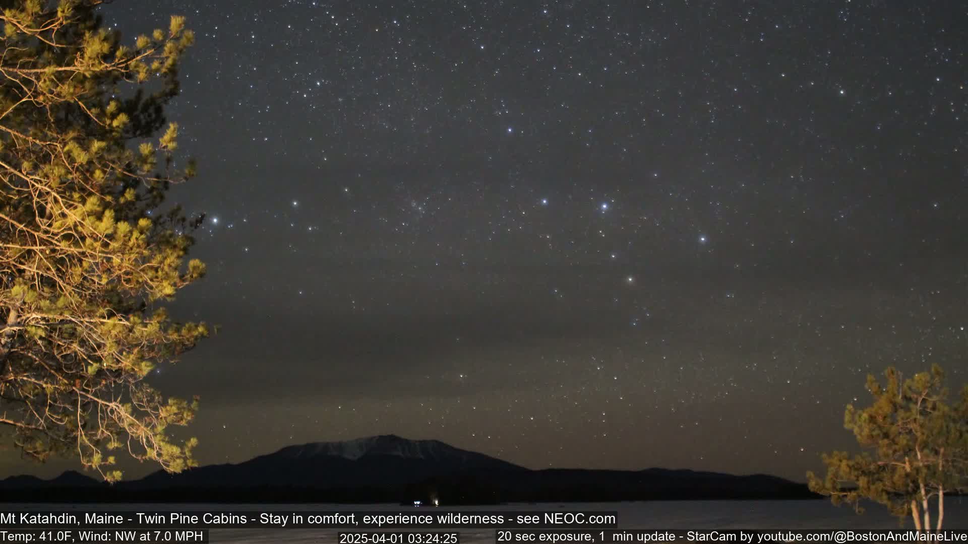 A nighttime view of a mountain range under a clear, star-filled sky with a calm body of water in the foreground, partially obscured by trees.