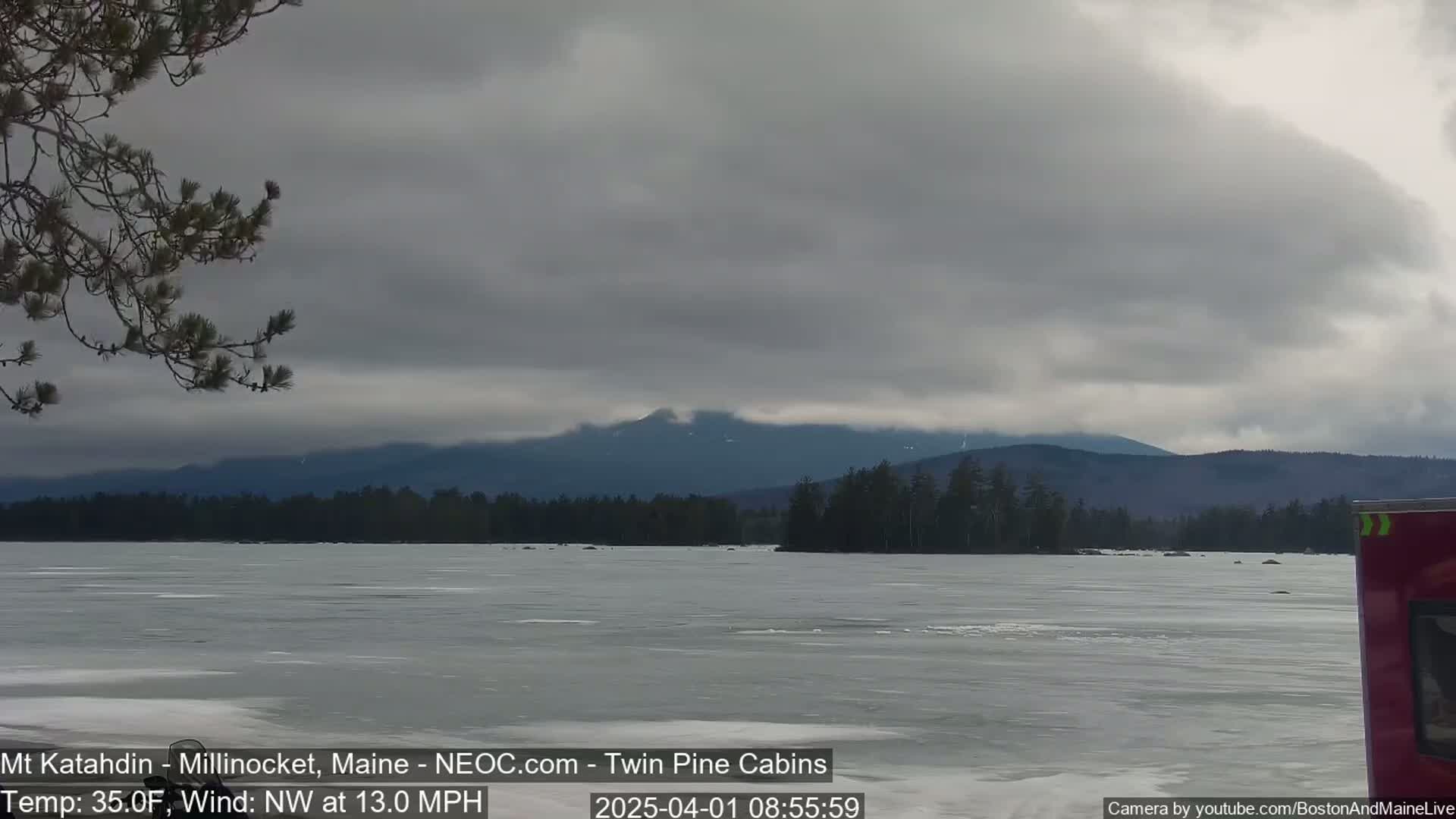 A frozen lake stretches towards a distant mountain range under a cloudy sky, with a northwest wind blowing at 13 miles per hour.