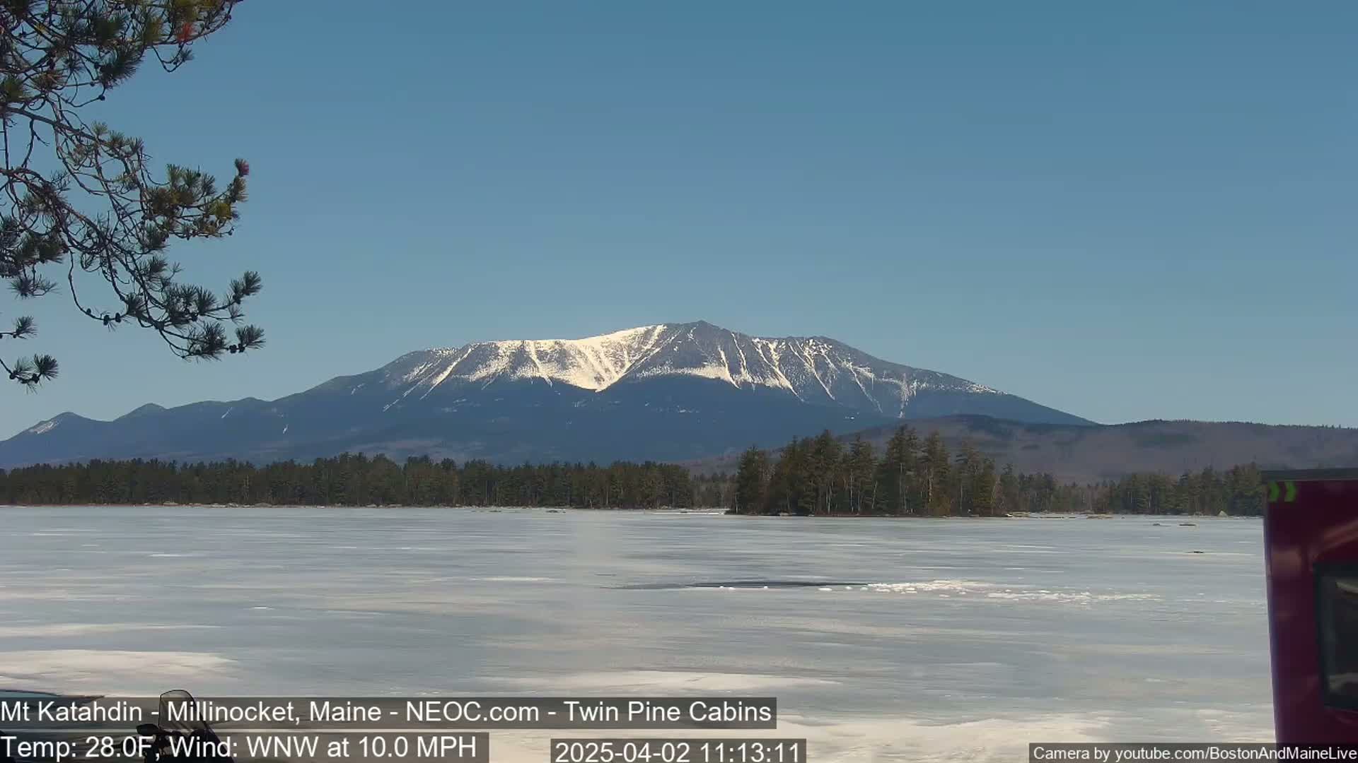 A snow-capped mountain is seen in the distance across a partially frozen lake under a clear blue sky.