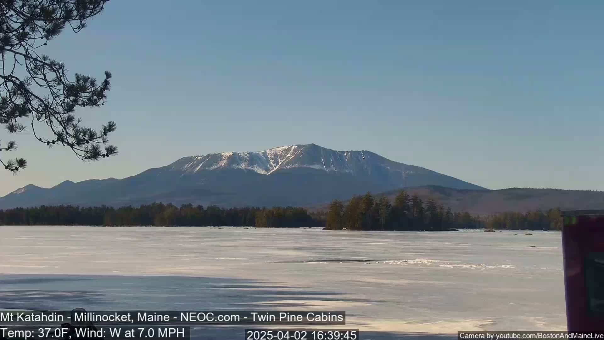 A partially frozen lake stretches out before a snow-capped mountain range under a clear, light blue sky.