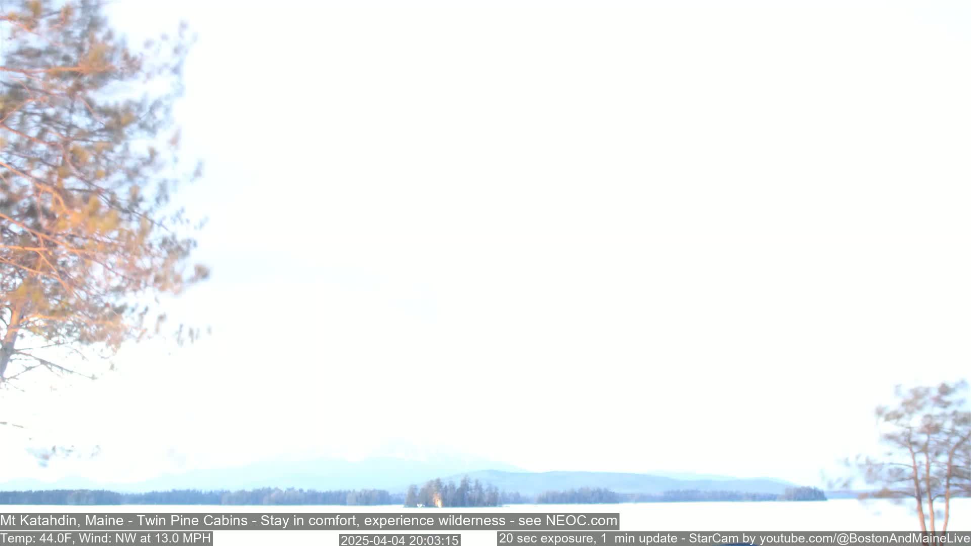 A snowy, hazy landscape with leafless trees in the foreground and a distant mountain range under a bright, white sky, with a northwest wind blowing at 13 mph.