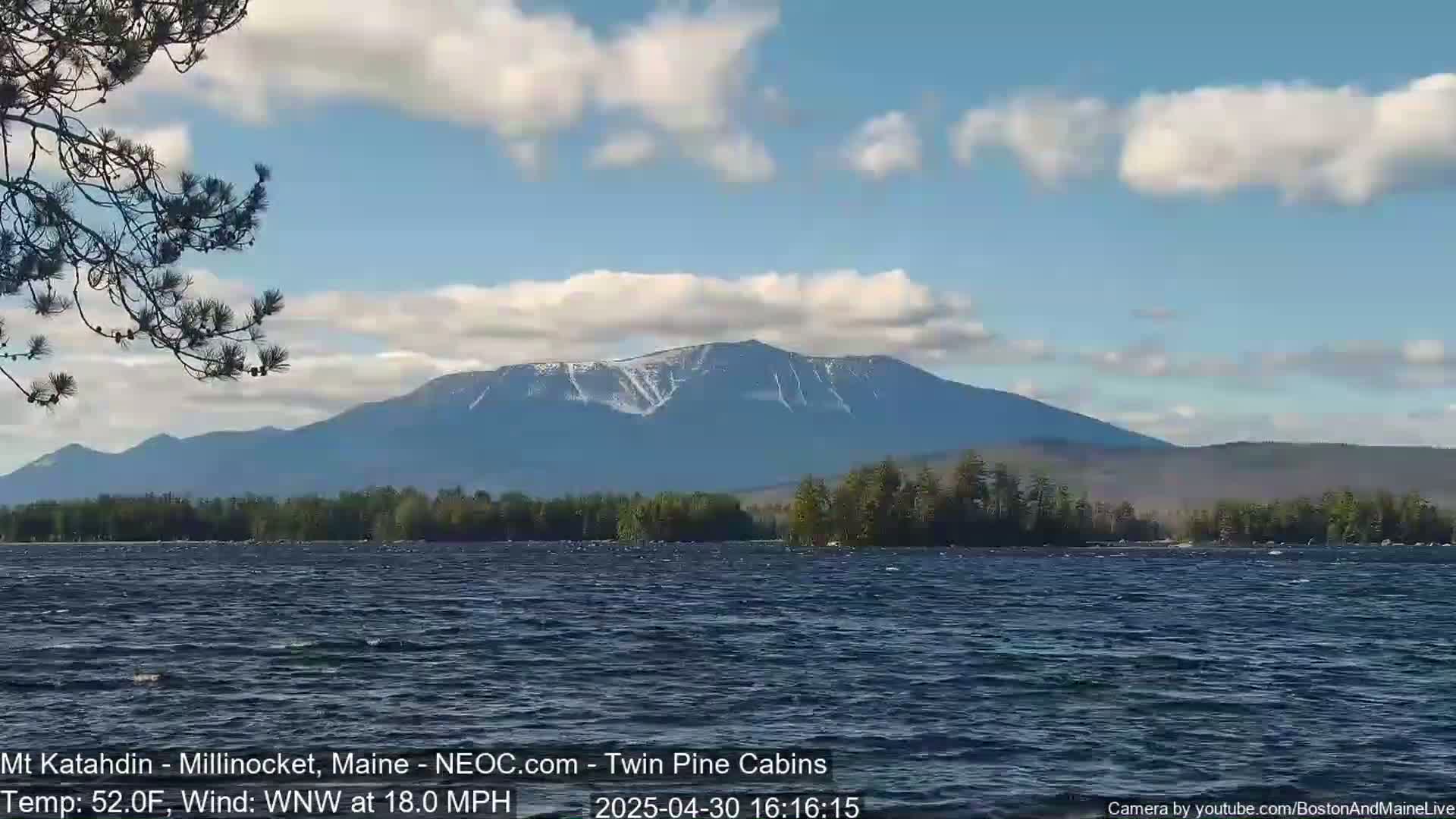 A partly cloudy sky overlooks a lake with a mountain range in the background, some snow visible on the peaks, and a moderate wind blowing.