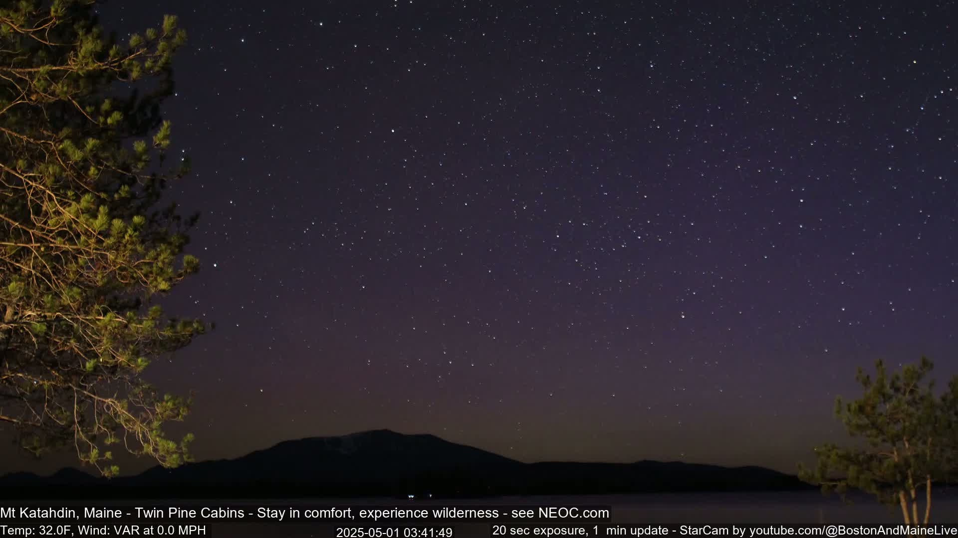 A night sky filled with stars is visible over a dark mountain range and silhouetted trees, with a calm, clear atmosphere.
