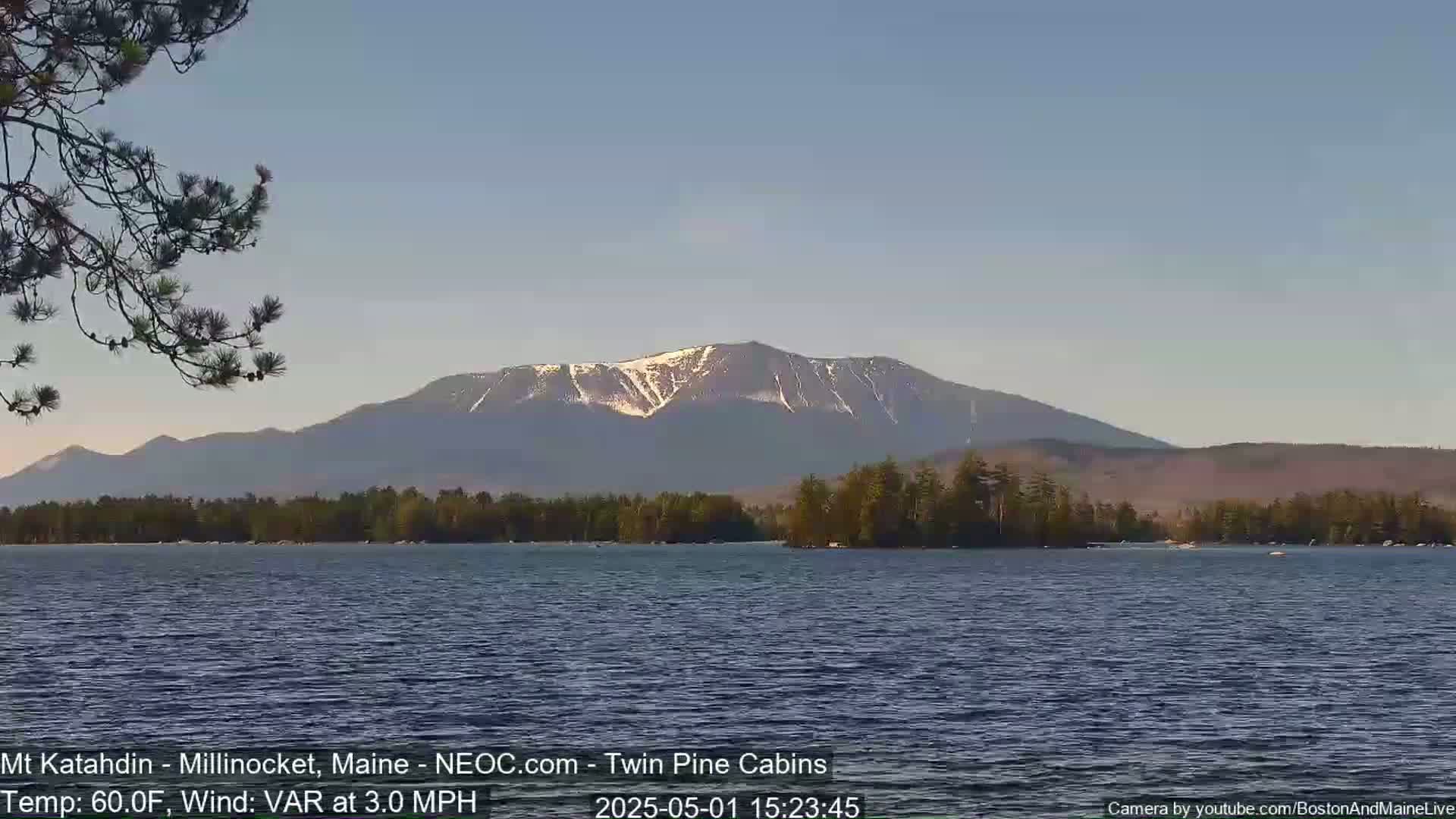 A calm lake reflects a mountain range with patches of snow under a clear, light-blue sky.